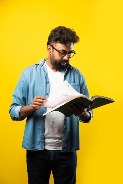 Giovane uomo indiano felice che indossa occhiali leggendo un libro e sorridendo isolato su giallo