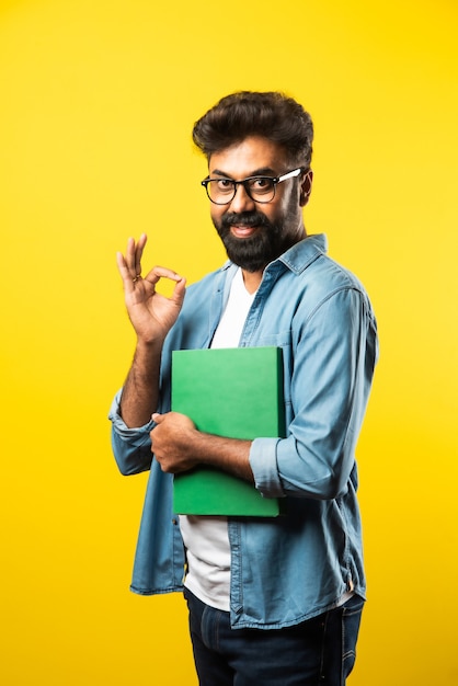 Young Indian happy man wearing eyeglasses reading book and smiling isolated on yellow
