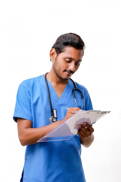 Молодой индийский мужчина-врач, одетый в униформу со стетоскопом, делая заметки в блокноте, изолированном на белом фоне.
