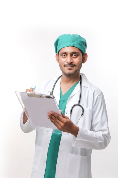 Giovane medico maschio indiano in uniforme con lo stetoscopio che prende le note in blocco note sopra fondo bianco.
