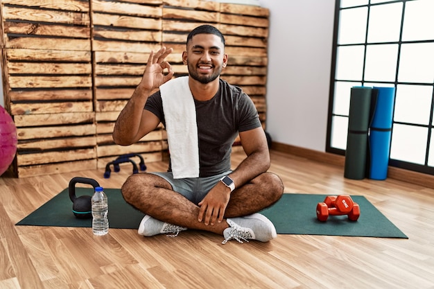 Young indian man sitting on training mat at the gym smiling positive doing ok sign with hand and fingers successful expression