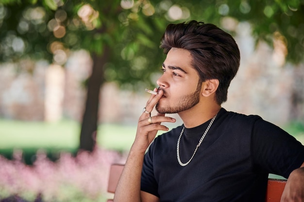 Young indian man smoker portrait in black t shirt and silver neck chain sitting on park bench