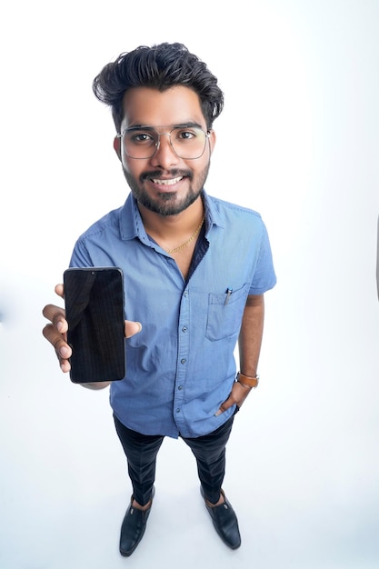 고립된 흰색 배경 위에 서 있는 스마트폰을 사용하는 젊은 인도 남자는 질문이나 솔루션에 대해 생각하는 심각한 얼굴을 하고 있습니다.