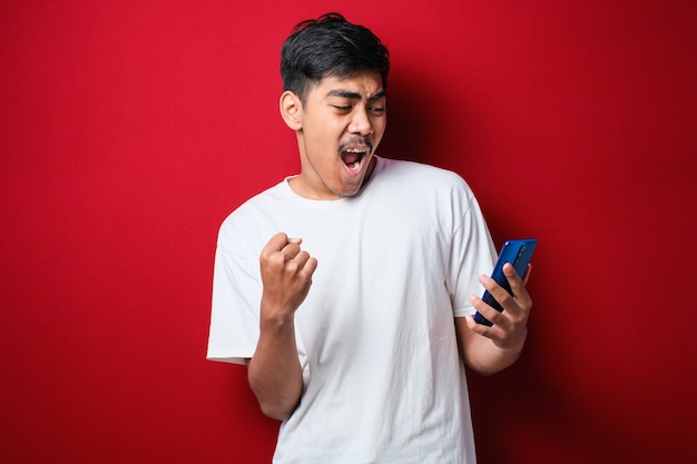 Young indian man using smartphone standing over isolated red background screaming proud and celebrating victory and success very excited, cheering emotion