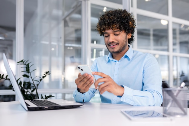 Young indian programmer inside modern office sitting at workplace man using phone testing online