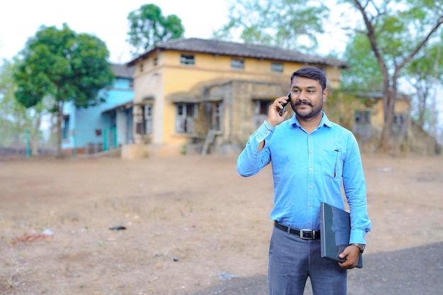 Young Indian property dealer standing in front of house with laptop talking on mobile phone