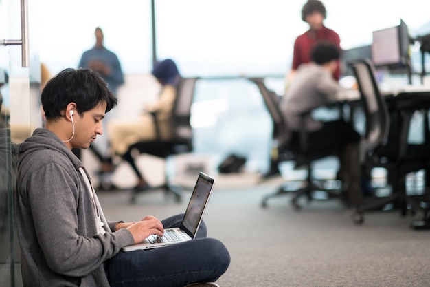 랩톱 컴퓨터를 사용하여 프로그래밍 코드를 작성하는 젊은 인도 소프트웨어 개발자 남자는 현대적인 크리에이티브 스타트업 사무실 바닥에 앉아 있습니다