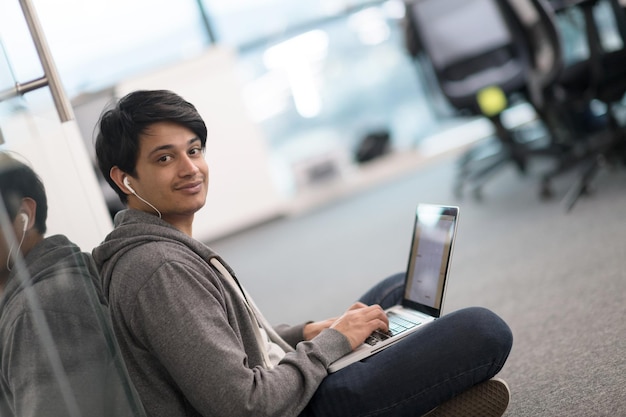 現代の創造的なスタートアップオフィスの床に座ってプログラミングコードを書くラップトップコンピューターを使用して若いインドのソフトウェア開発者の男