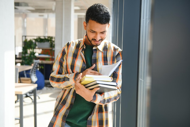 Young indian student boy reading book studying in college library with bookshelf behind working on assignment or project