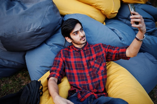 Young indian student man at checkered shirt and jeans sitting and relax at outdoor pillows and making selfie