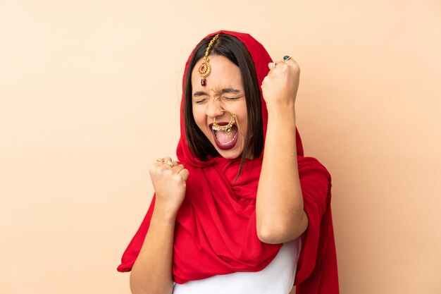 Young Indian woman on beige wall celebrating a victory