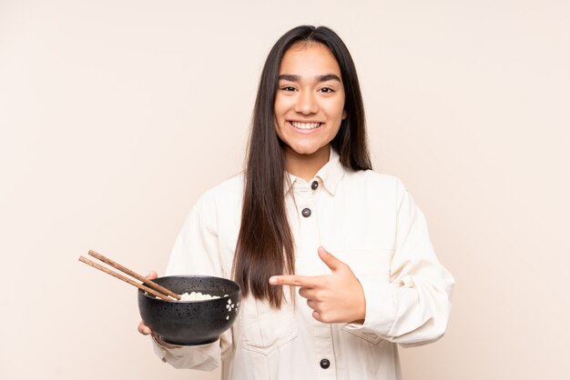 Young Indian woman on beige wall and pointing it while holding a bowl of noodles with chopsticks