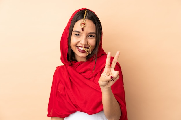 Young Indian woman on beige wall smiling and showing victory sign