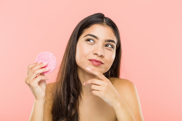 Young indian woman holding a facial skin care disc looking sideways with doubtful and skeptical expression.