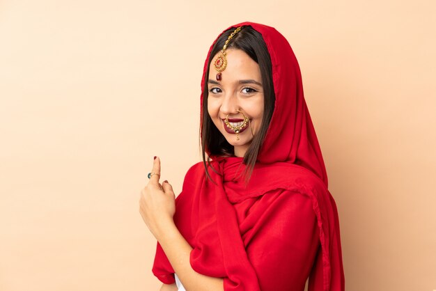 Young Indian woman isolated on beige wall pointing back