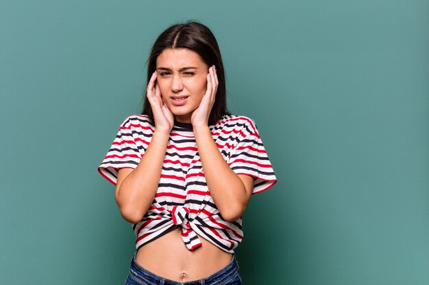 Young Indian woman isolated on blue background covering ears with fingers, stressed and desperate by a loudly ambient.