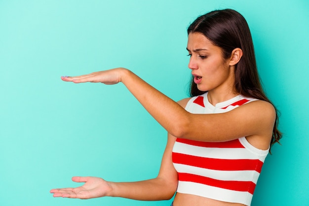 Young Indian woman isolated on blue background shocked and amazed holding a copy space between hands.