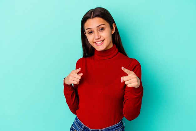 Young Indian woman isolated on blue wall cheerful smiles pointing to front.