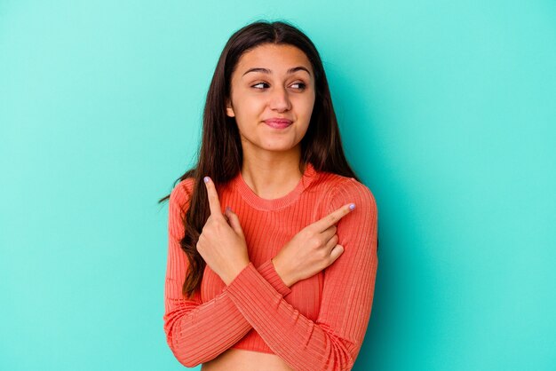 Young Indian woman isolated on blue wall points sideways, is trying to choose between two options.