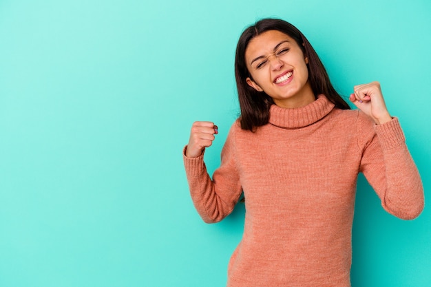 Young Indian woman isolated on blue wall raising fist after a victory, winner concept.