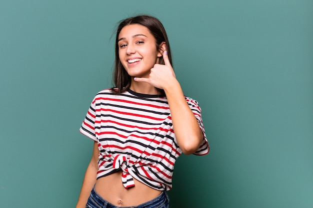 Young Indian woman isolated on blue wall showing a mobile phone call gesture with fingers.