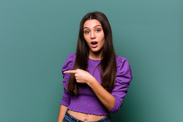 Young Indian woman isolated on blue wall smiling and pointing aside, showing something at blank space.