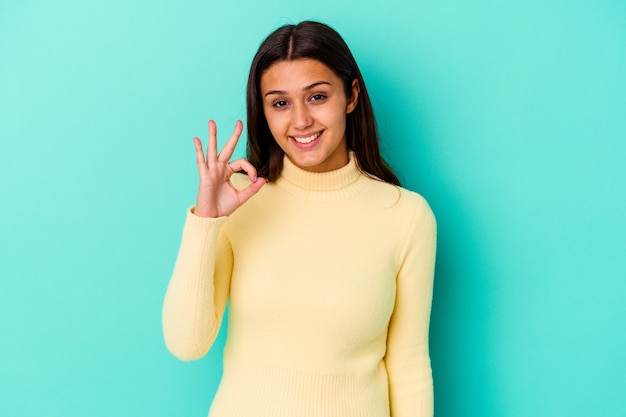 Young Indian woman isolated on blue wall winks an eye and holds an okay gesture with hand.