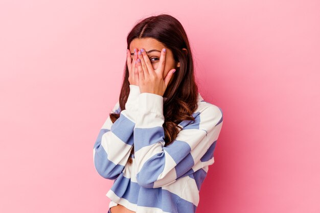 Young Indian woman isolated on pink background blink through fingers frightened and nervous.