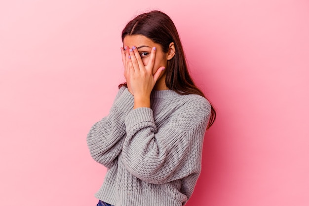 Young Indian woman isolated on pink background blink through fingers frightened and nervous.