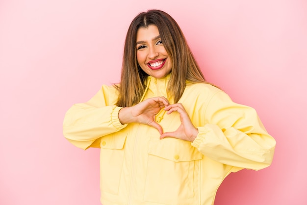 Young indian woman isolated on pink background smiling and showing a heart shape with hands.