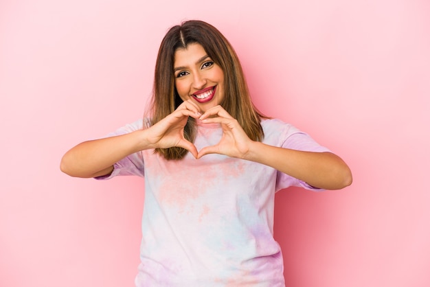 Young indian woman isolated on pink background smiling and showing a heart shape with hands.