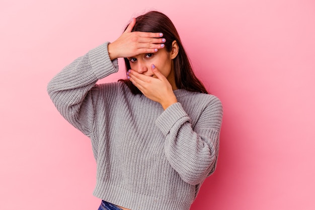 Young Indian woman isolated on pink wall blink through fingers, embarrassed covering face.