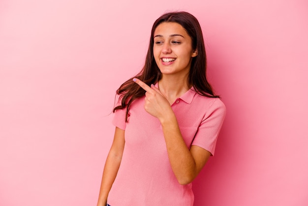 Young Indian woman isolated on pink wall looks aside smiling, cheerful and pleasant.