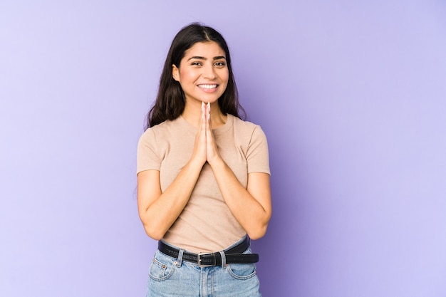 Young indian woman isolated on purple background holding hands in pray near mouth, feels confident.