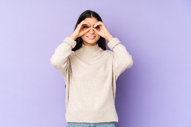 Young indian woman isolated on purple wall showing okay sign over eyes