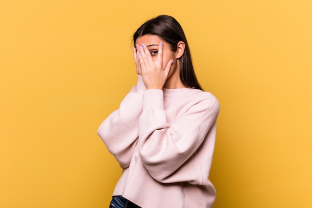 Young Indian woman isolated on yellow wall blink through fingers frightened and nervous.