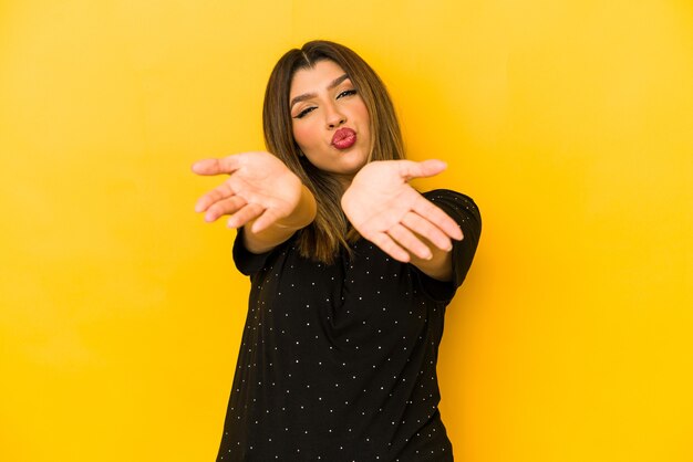 Giovane donna indiana isolata sul muro giallo, piegando le labbra e tenendo i palmi delle mani per inviare un bacio d'aria.