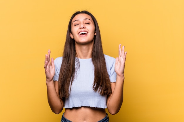 Young Indian woman isolated on yellow wall laughs out loudly keeping hand on chest.