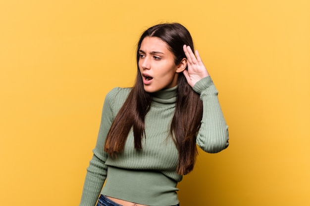 young indian woman isolated on yellow wall trying to listening a gossip