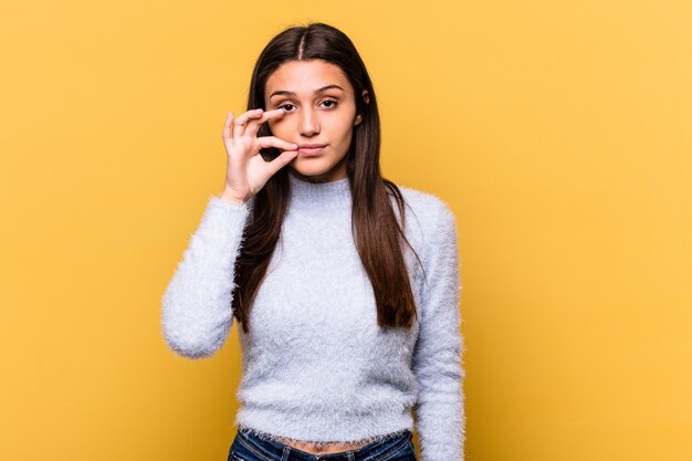 Young Indian woman isolated on yellow wall with fingers on lips keeping a secret.