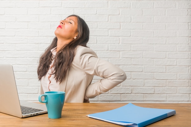 Young indian woman at the office with back pain due to work stress, tired and astute