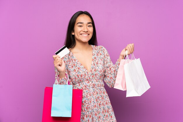 Young Indian woman on purple wall holding shopping bags and a credit card