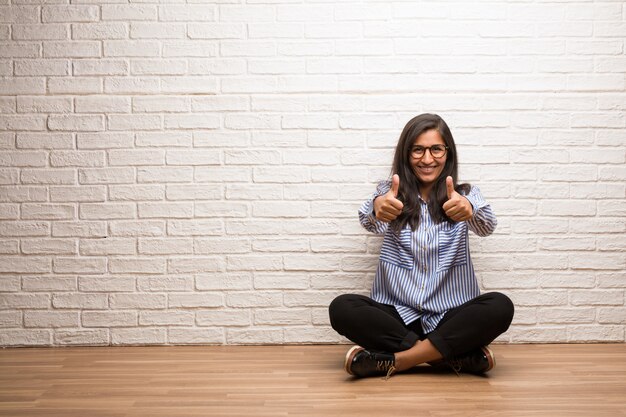 Молодая индийская женщина сидит у кирпичной стены, веселая и взволнованная, улыбаясь и поднимая ее