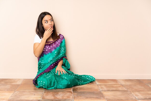 Young Indian woman sitting on the floor doing surprise gesture while looking to the side