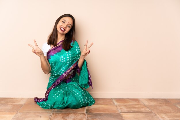 Foto giovane donna indiana che si siede sul pavimento che mostra il segno di vittoria con entrambe le mani