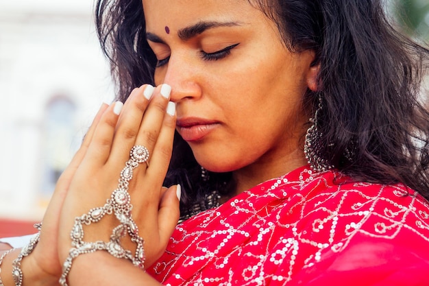 Giovane donna indiana nel tradizionale sari vestito rosso che prega in un tempio indù goa india hinduismgirl che esegue il gesto di namaste cattolicesimo delhi street holi festivalom meditazione yoga modello femminile