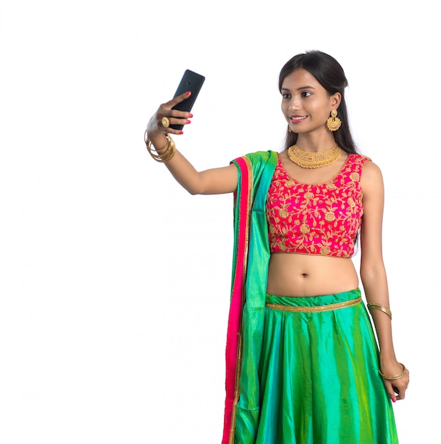 Giovane donna indiana che per mezzo di un telefono cellulare o di uno smartphone, parlando selfie o parlando in video chat isolato su un muro bianco