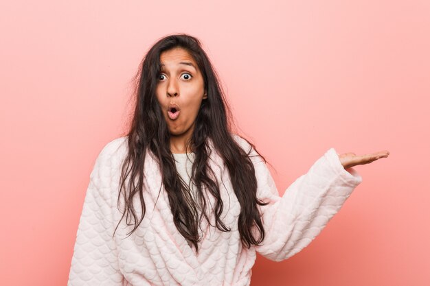 Young indian woman wearing pajama impressed holding on palm.