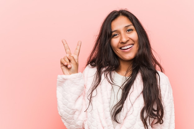 Young indian woman wearing pajama joyful and carefree showingpeace symbol with fingers.