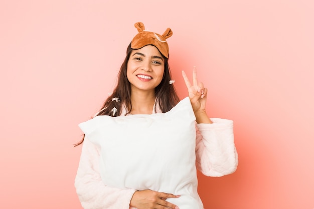 Young indian woman wearing a pajamas and sleep mask isolated holding a pillow young indian woman wearing a pajamas and sleep mask isolated showing victory sign and smiling broadly.< mixto >